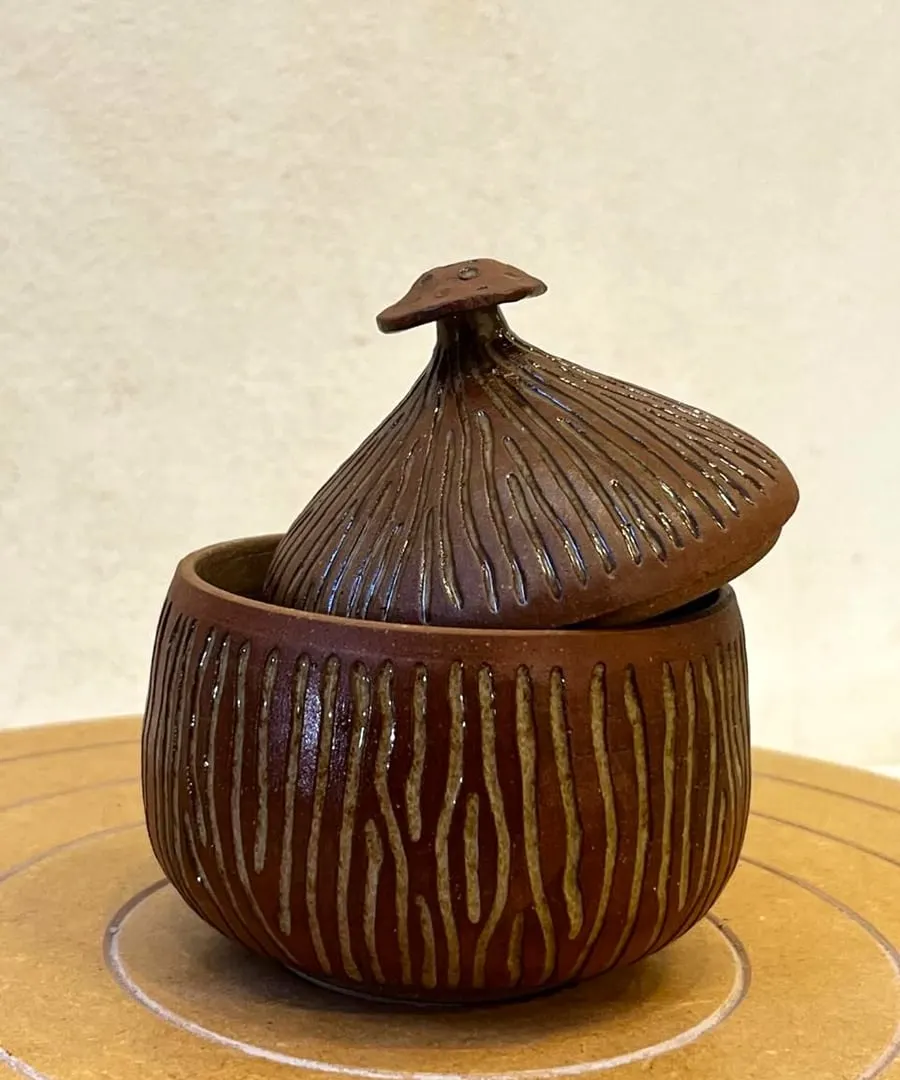 Tarro marrón con tapa en forma de hongo y líneas verticales talladas.